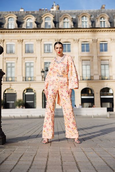 Salopeta SOLARIA FLOWERS cu maneci lungi, cu un decolteu in forma de V inconjurat de cristale, cu pantaloni lungi si largi, din satin portocaliu deschis peste care am aplicat o dantela din bumbac multicolora, cu cordon in talie